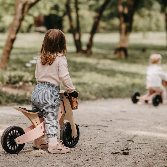 Image is of child standing with two wheeled wooden pink bike on a path with child on wooden tricycle ahead. Children are not shown wearing helmets. Helmets are strongly recommended and required by law. 