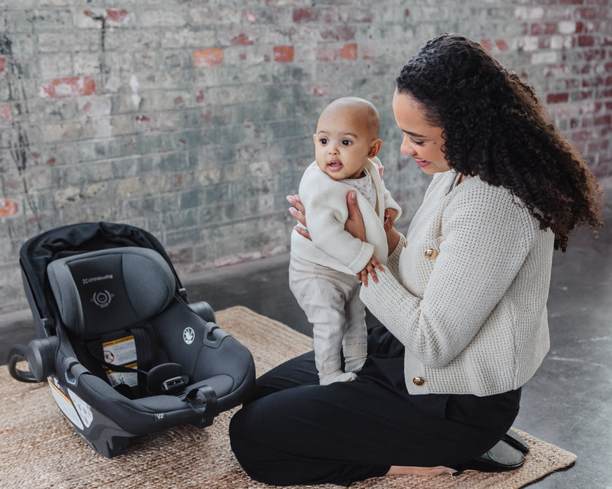 Woman holding a baby next to an uppababy Aria V2car seat in an urban setting