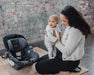 Woman holding a baby next to an uppababy Aria V2car seat in an urban setting