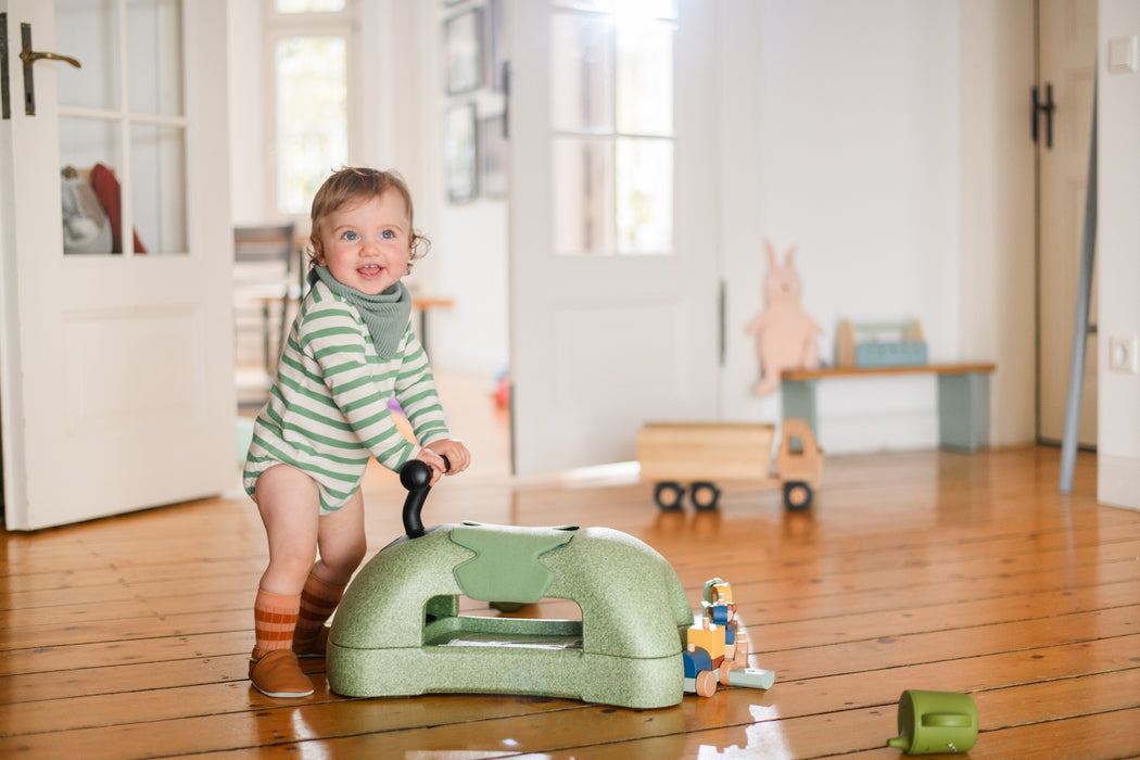 Child standing next to green ride on toy in living room setting
