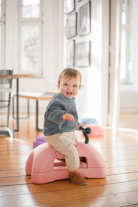 Child sitting on a pink toy ride on toy in a bright room with large windows.