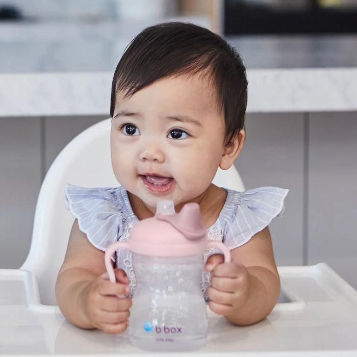 Baby sitting in a high chair holding a pink sippy cup with a white background