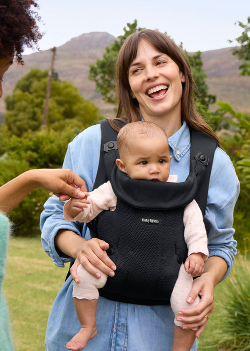 A woman outdoors wearing a black baby carrier with a baby facing out in front of her. The carrier has a breathable mesh front panel and padded shoulder straps.
