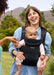 A woman outdoors wearing a black baby carrier with a baby facing out in front of her. The carrier has a breathable mesh front panel and padded shoulder straps.