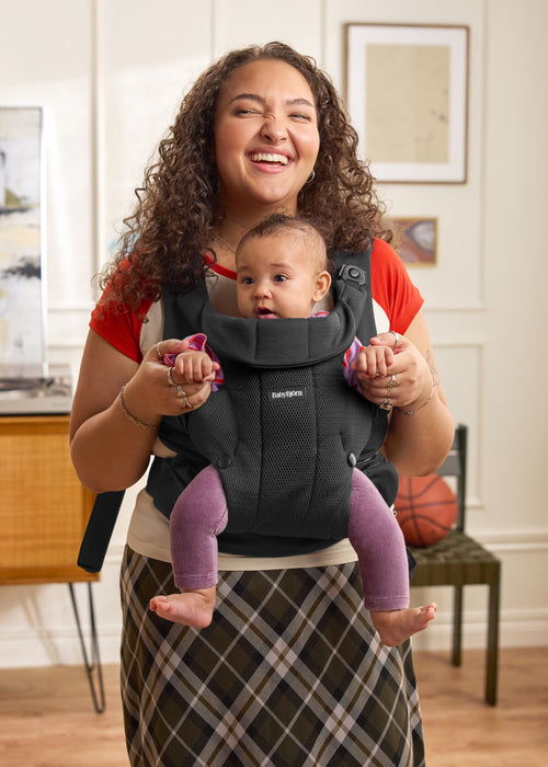 A woman wearing a red shirt and a plaid skirt holds a baby in a black Baby Bjorn baby carrier. The baby is wearing purple leggings.