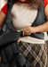 Close-up of a person putting on a black mesh Baby Bjorn Hold carrier like a vest over a cream top and plaid skirt, fastening the carrier with a buckle.