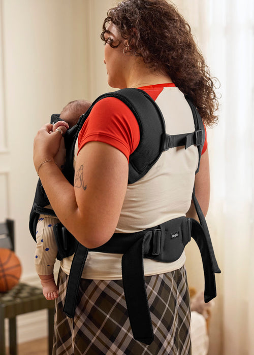 A woman wearing a black baby carrier holding a baby, shown from the back for fit details in an indoor setting.