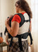 A woman wearing a black baby carrier holding a baby, shown from the back for fit details in an indoor setting.