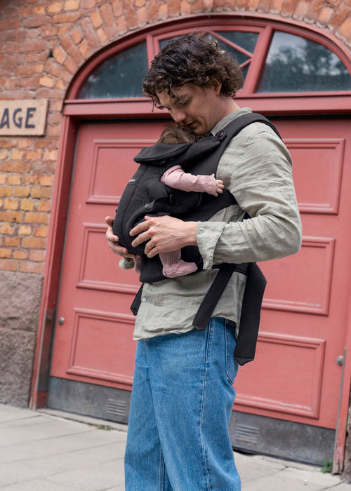 A man wearing a light gray long-sleeve shirt and blue jeans carries a baby in a black baby carrier in front of a red arched door.