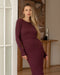 Woman leans against a doorframe wearing a long sleeve burgundy maternity and nursing dress in a ribbed fabric