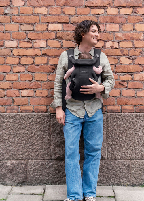 A man wearing a black baby carrier with a baby wearing a pink outfit, standing against a red brick wall.