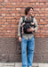 A man wearing a black baby carrier with a baby wearing a pink outfit, standing against a red brick wall.