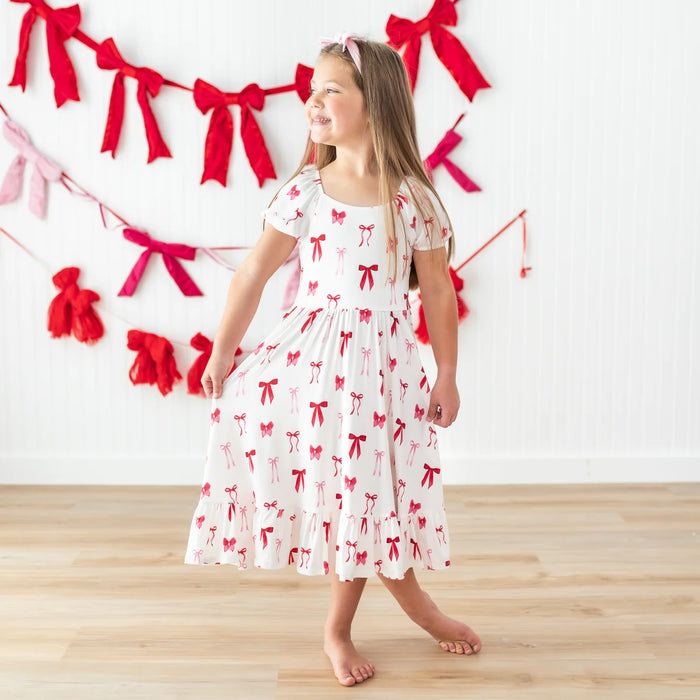 Young girl in a white dress with red bow pattern standing in front of a decorative wall with red bows.