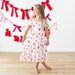Young girl in a white dress with red bow pattern standing in front of a decorative wall with red bows.