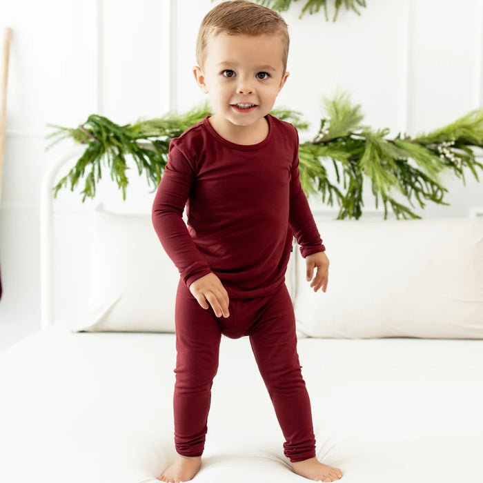Child wearing a burgundy long-sleeve pj shirt and pants inside with tree trimming in the background