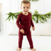 Child wearing a burgundy long-sleeve pj shirt and pants inside with tree trimming in the background