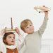 Two children playing with wooden toy vehicles against a white wall.