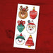 A sheet of Christmas-themed stickers featuring a reindeer head, patterned ornaments, Santa Claus, a snowman, and a bell, displayed on a red wooden background.