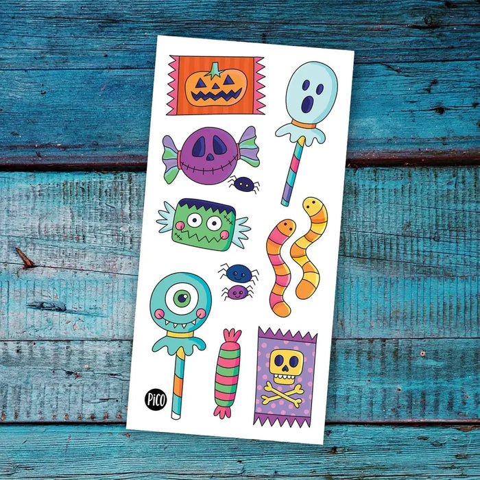 A sheet of colorful Halloween-themed temporary tattoos or stickers displayed flat on a rustic, weathered blue wooden surface. The sheet features cartoon illustrations of a jack-o'-lantern, ghost lollipop, purple skull candy, green Frankenstein monster, orange and yellow striped snake, and a skull-and-crossbones on a purple flag.