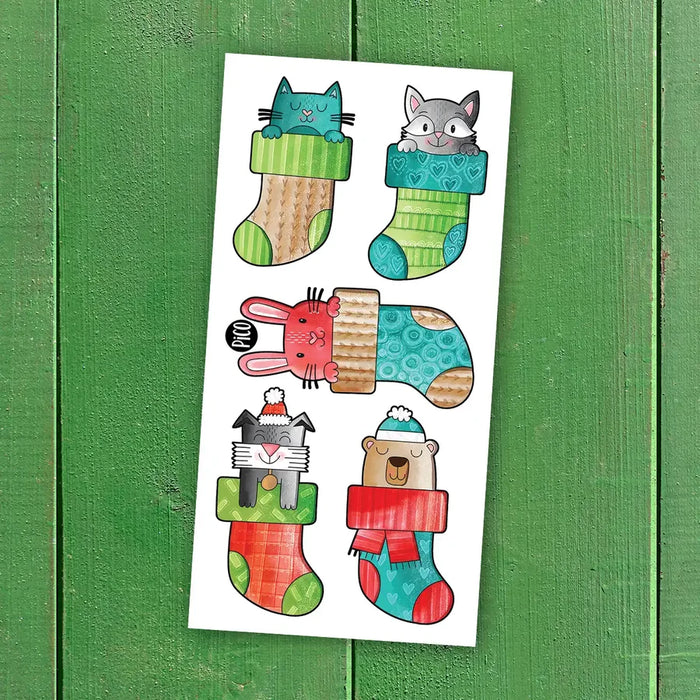 A sheet of cartoon animal Christmas stockings featuring a blue cat, gray raccoon, pink bunny, gray cat, and a brown bear peeking out of colorful Christmas socks, displayed on a green wooden background.