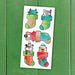 A sheet of cartoon animal Christmas stockings featuring a blue cat, gray raccoon, pink bunny, gray cat, and a brown bear peeking out of colorful Christmas socks, displayed on a green wooden background.