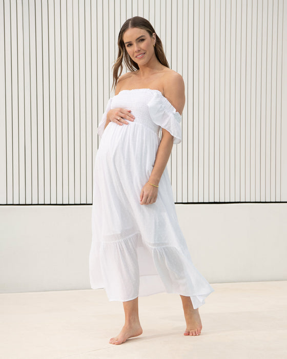 Woman wearing a white off-shoulder maternity dress against a minimalistic background