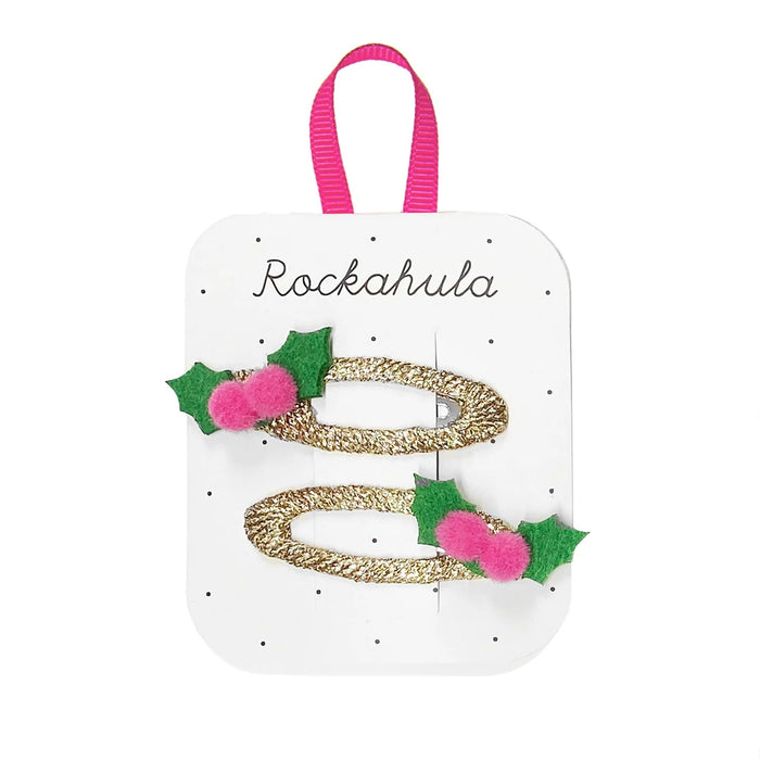 Two gold glitter hair clips decorated with green felt holly leaves and pink pom-poms, attached to a white display card with a pink ribbon loop and the brand name 'Rockahula'.