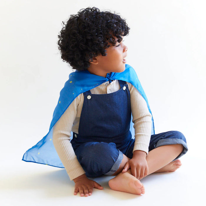 Child wearing a blue silk as a cape sitting on a white floor