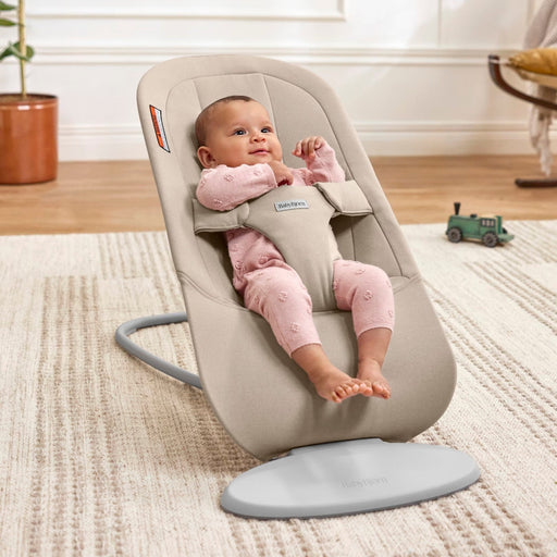 A baby in a pink outfit sits in a beige baby bouncer chair on a textured cream rug in a bright room with a wooden floor, potted plant, and toy car.