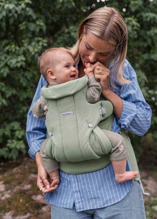 A woman wearing a blue and white striped shirt carries a baby in a sage green Baby Bjorn baby carrier outdoors against a green foliage background.