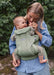 A woman wearing a blue and white striped shirt carries a baby in a sage green Baby Bjorn baby carrier outdoors against a green foliage background.