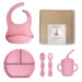 Pink baby feeding set with bib, plate, spoons, and cup on a white background