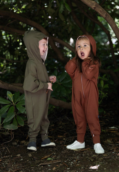 Two children wearing hooded one-piece jumpsuits standing outdoors in a wooded area. The child on the left wears an olive green jumpsuit, and the child on the right wears a rust brown jumpsuit.