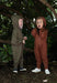 Two children wearing hooded one-piece jumpsuits standing outdoors in a wooded area. The child on the left wears an olive green jumpsuit, and the child on the right wears a rust brown jumpsuit.