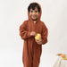 A young child wearing a rust-brown hooded zip-up onesie, holding a yellow apple against a clean white studio background.