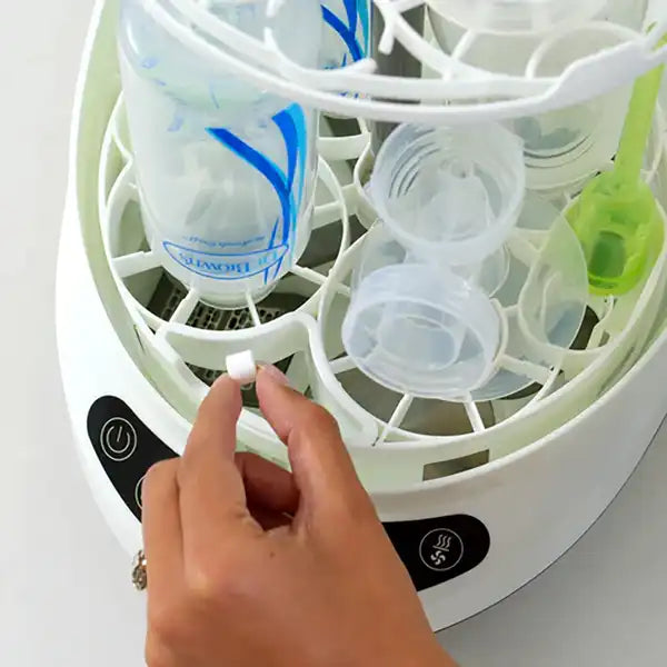 Hand placing a detergent tablet into a white bottle sterilizer with bottles inside.
