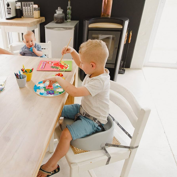 Two children sitting at a table with art supplies, one using a bumbo booster seat.