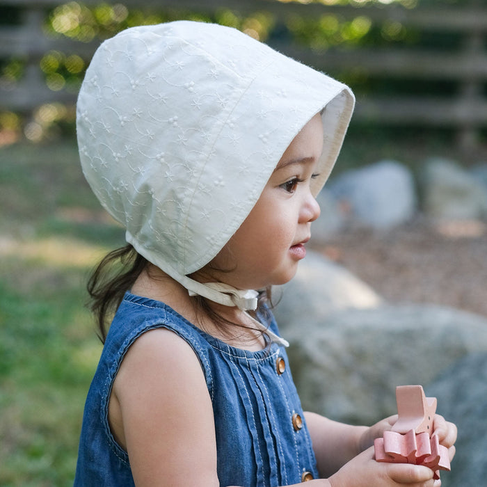 Child wearing a white bonnet and blue dress, holding a pink object outdoors.