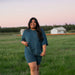 Woman in teal outfit standing in a grassy field with a sunset background