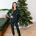 Woman wearing a black kyte pajama set with green tree pattern in a room with a Christmas tree.