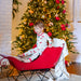 Child in kyte pajamas sitting in a red sleigh in front of a decorated Christmas tree.