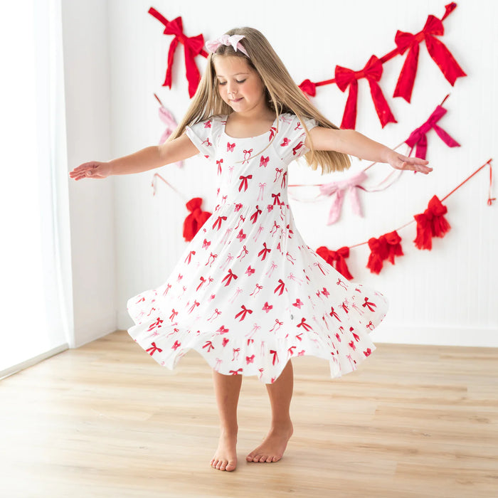 Young girl in a white bamboo dress with red bows twirling in front of a decorative bow banner.