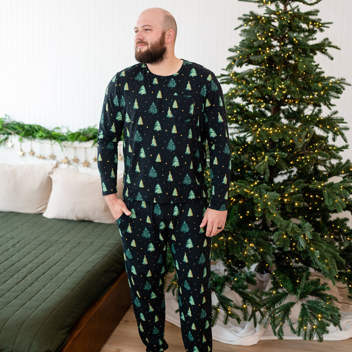 Man wearing a black Christmas tree pattern kyte pajama set standing in front of a decorated Christmas tree.