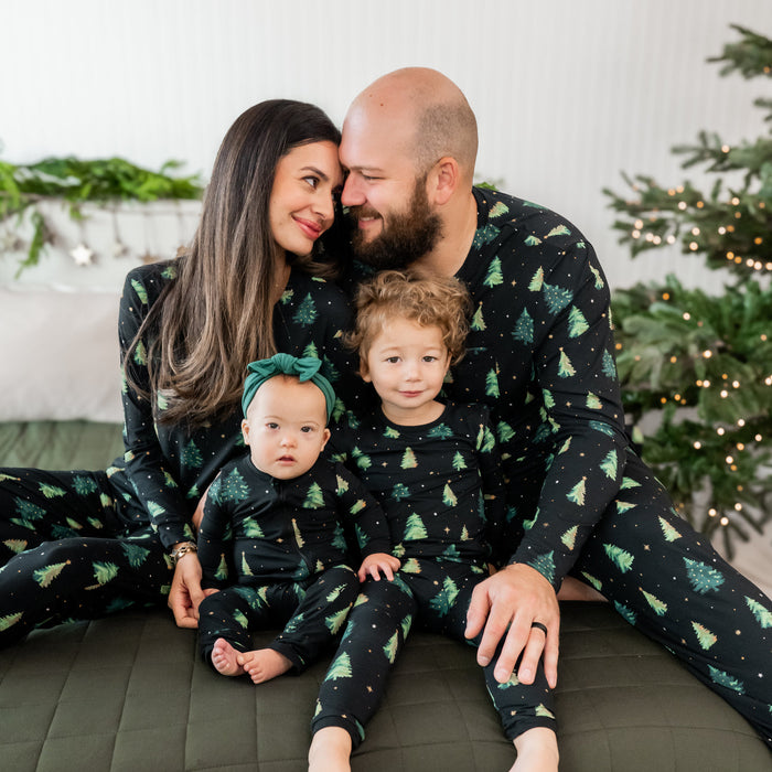 Family of four wearing matching black kyte pajamas with green tree patterns, sitting on a couch in a festive setting.