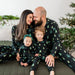 Family of four wearing matching black kyte pajamas with green tree patterns, sitting on a couch in a festive setting.