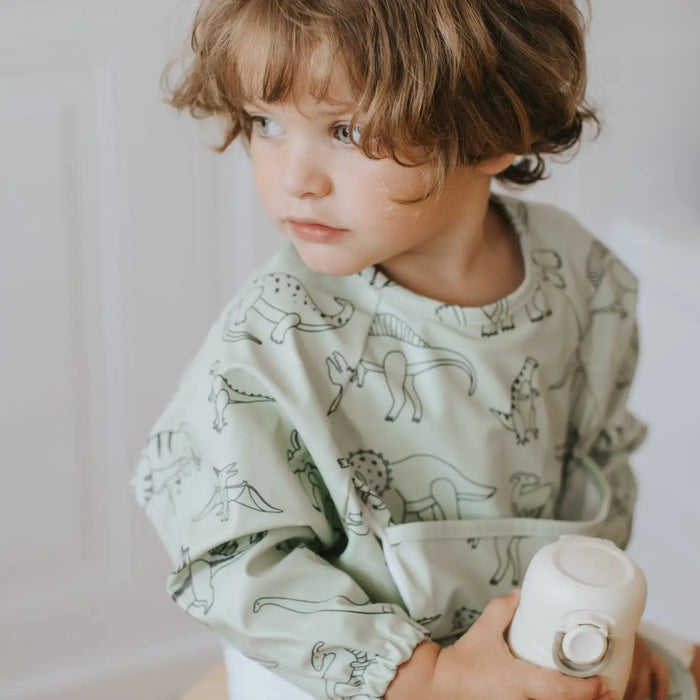 A toddler with curly brown hair wearing a sage green long-sleeve bib with a black dinosaur pattern, holding a white bottle in a brightly lit indoor setting.