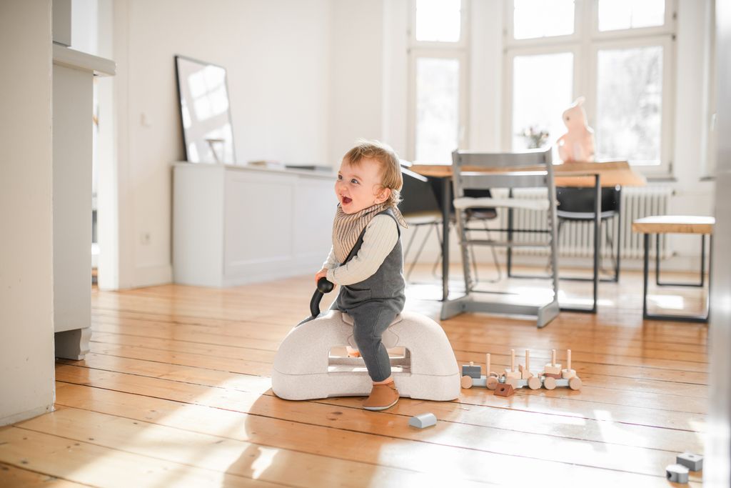 Child sitting on a white stool in a modern room with wooden floor and large windows.