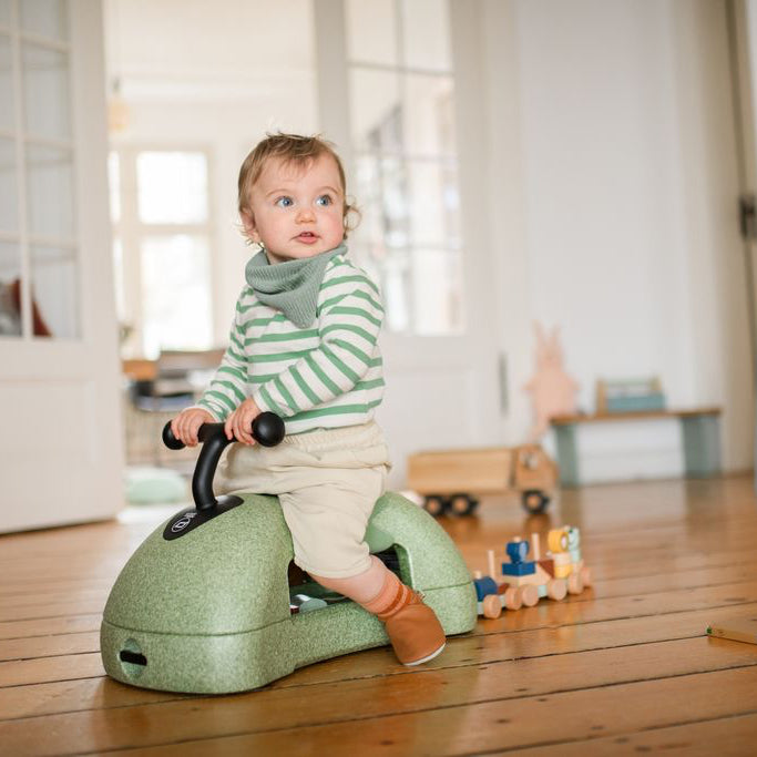 Child riding a green toy horse indoors on a wooden floor.
