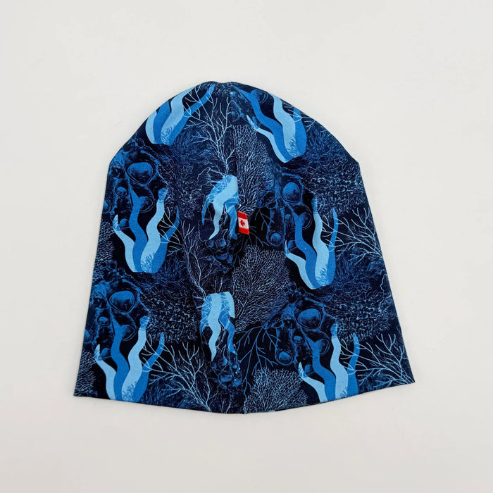 A blue patterned beanie hat featuring a dark navy background with light blue ocean figures and coral-like illustrations, laid flat on a light off-white background.
