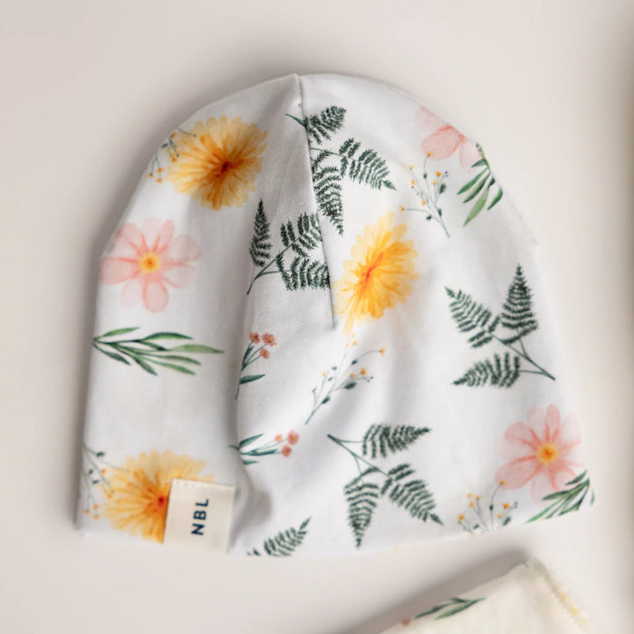 A baby beanie hat with a white background and a colorful floral and fern pattern, featuring yellow flowers, pink blossoms, and green leaves. A small fabric tag on the brim reads 'NBL'.